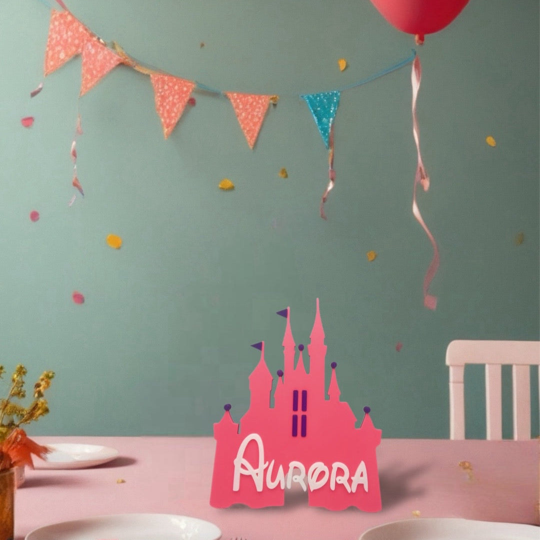 A bright pink, 3D-printed castle silhouette with the name "AURORA" cut out in white, standing upright on a pink tablecloth. The castle has turrets, flags, and window details. The background shows a teal wall with a colourful banner, a pink balloon, plates, and cutlery, suggesting a party setting.