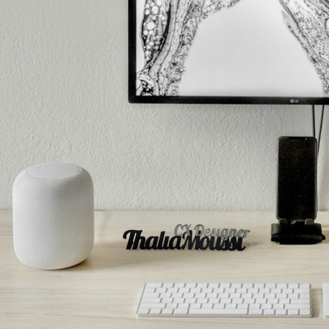 A black 3D-printed desk nameplate that reads "Thalia Moussi" with "CX Designer" above it, sitting on a light wooden desk in a modern workspace. A white cylindrical speaker is to the left, a black smartphone in a dock is behind it, and a white keyboard is in the foreground, beneath a computer monitor displaying a black and white abstract image.