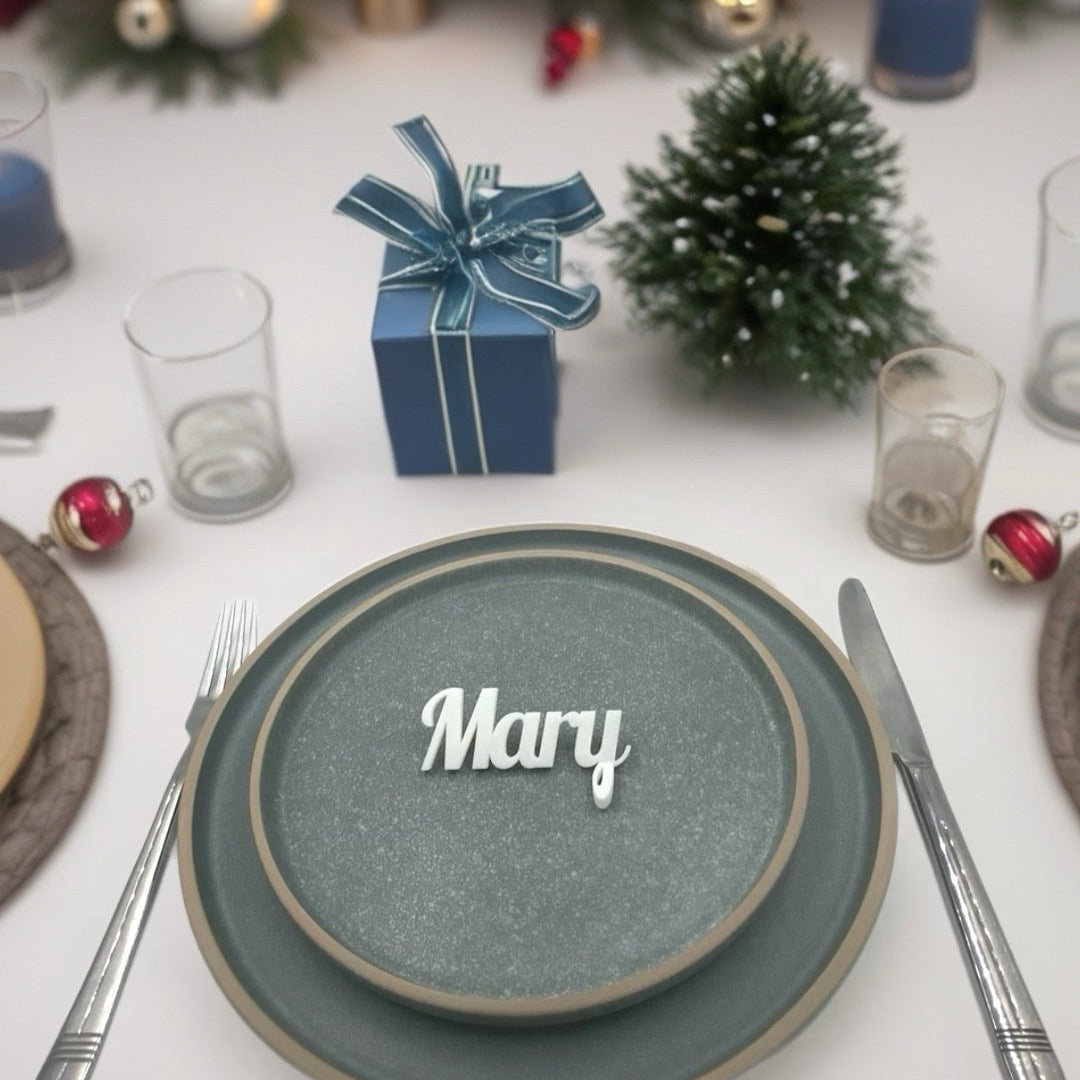 
A white 3D printed personalised place name "Mary" displayed on a textured grey plate in a festive table setting with Christmas decorations.

