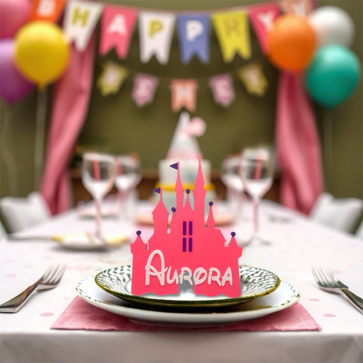 
A bright pink, castle-shaped place card with the name "Aurora" in white, sits on a decorative plate at a party table. In the blurred background, colourful balloons and a "Happy Birthday" banner are visible.