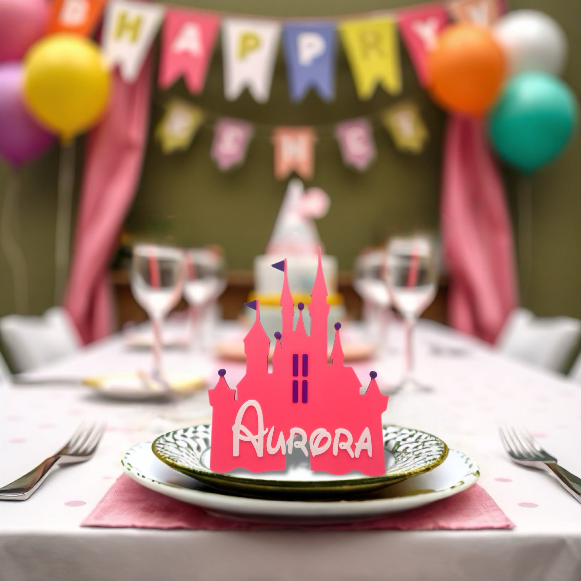 
A bright pink, castle-shaped place card with the name "Aurora" in white, sits on a decorative plate at a party table. In the blurred background, colourful balloons and a "Happy Birthday" banner are visible.