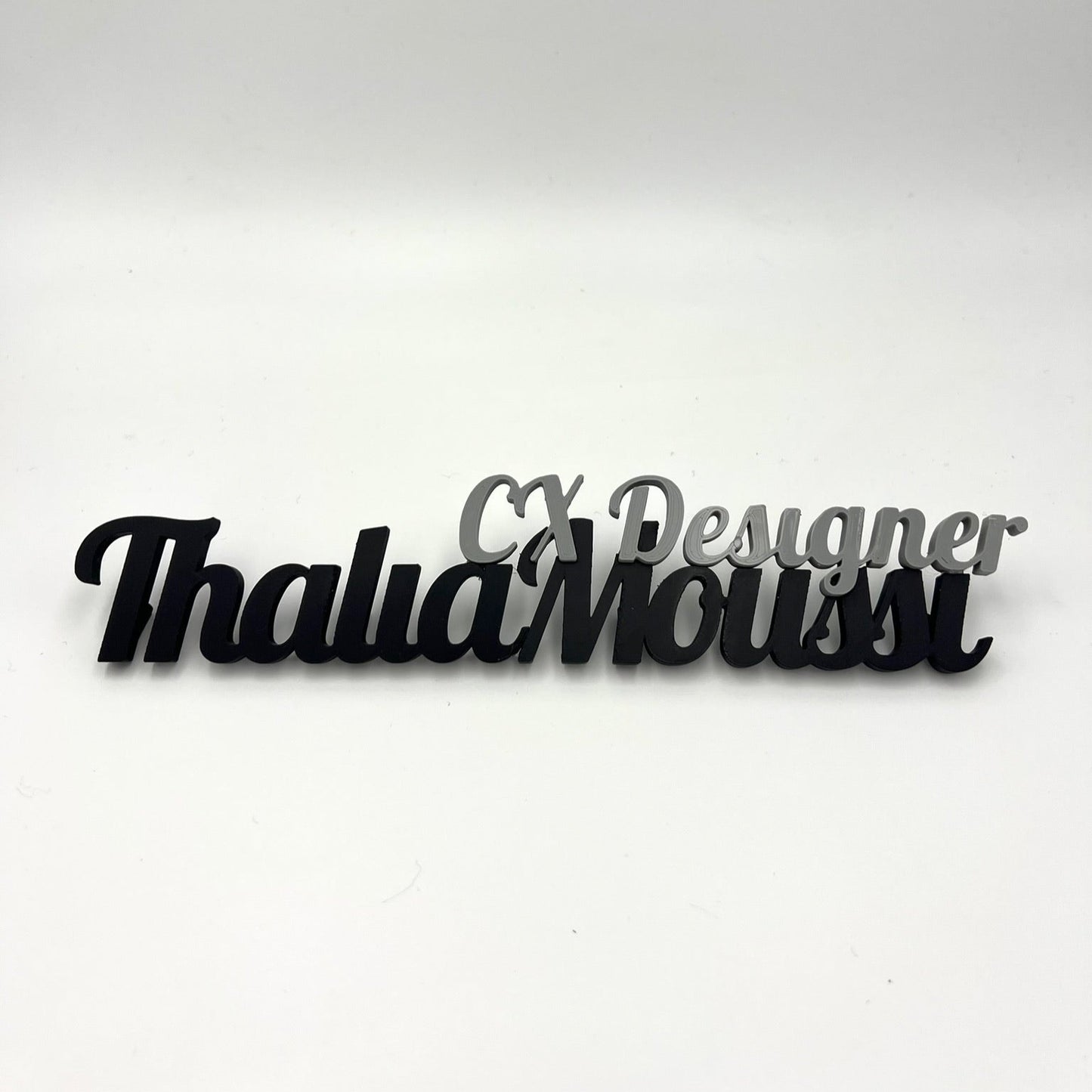 A black and grey 3D-printed desk nameplate that says "Thalia Moussi" with "CX Designer" above it, on a white background.
