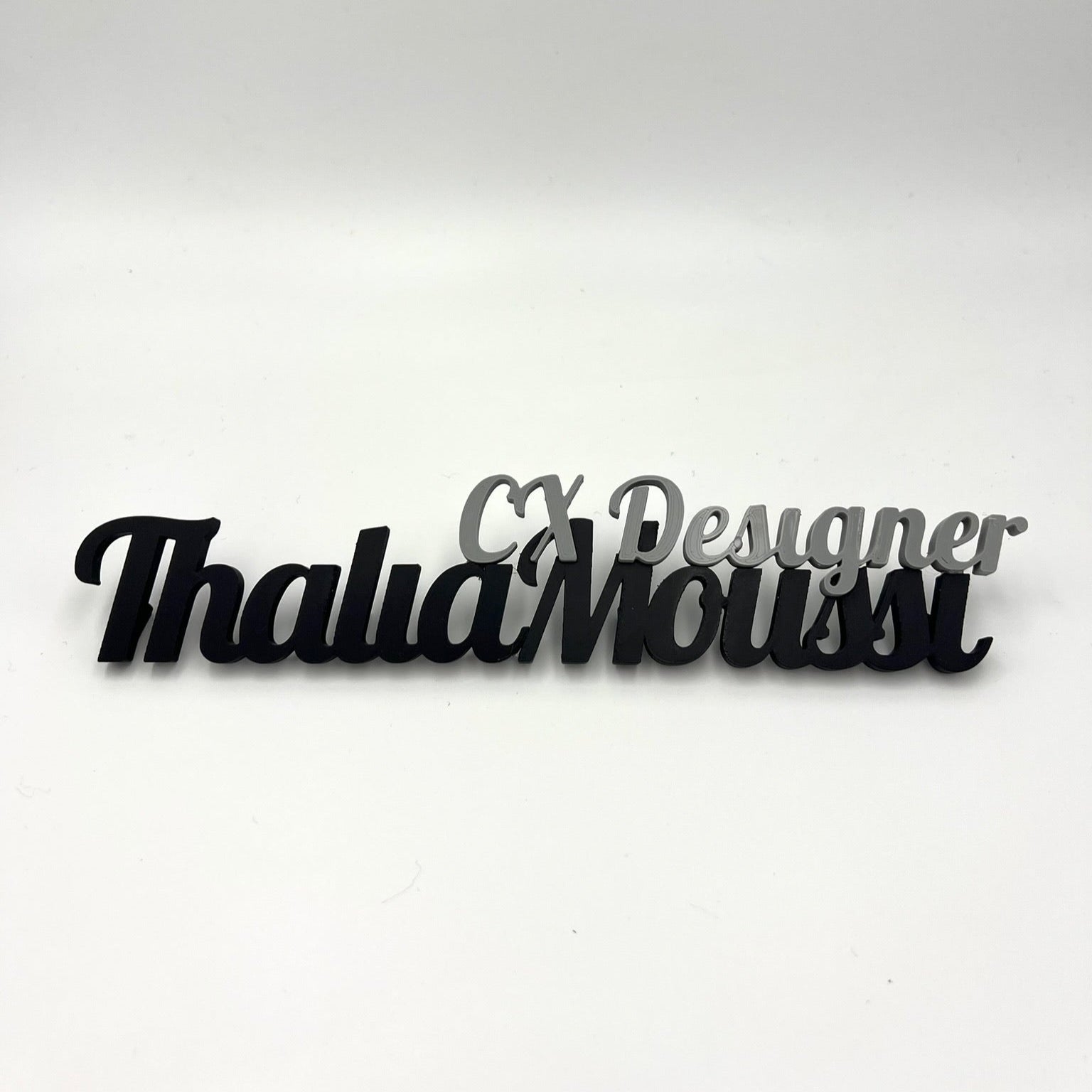 A black and grey 3D-printed desk nameplate that says "Thalia Moussi" with "CX Designer" above it, on a white background.
