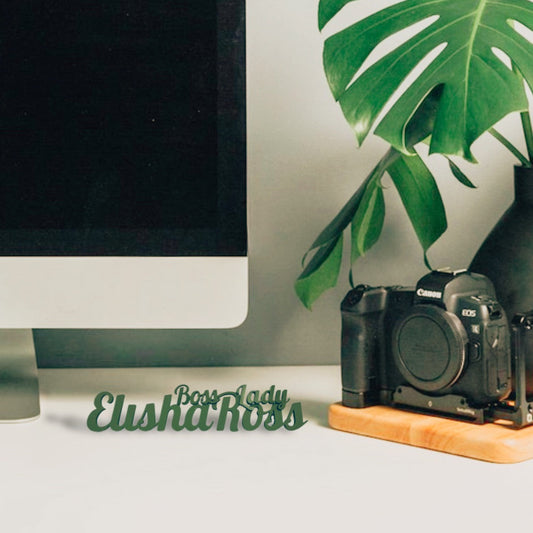 A green 3D-printed desk nameplate that says "Elisha Ross" with "Boss Lady" above it, sitting on a white desk with a computer, camera, and plant in the background.