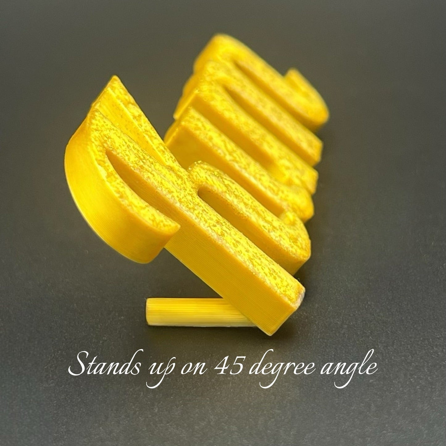 An angled close-up of a gold glitter 3D printed personalised name plate spelling "Kim" standing upright at approximately a 45-degree angle on a dark grey surface. The integrated stand supporting the nameplate is visible beneath the "K." Text at the bottom of the image reads "Stands up on 45 degree angle."