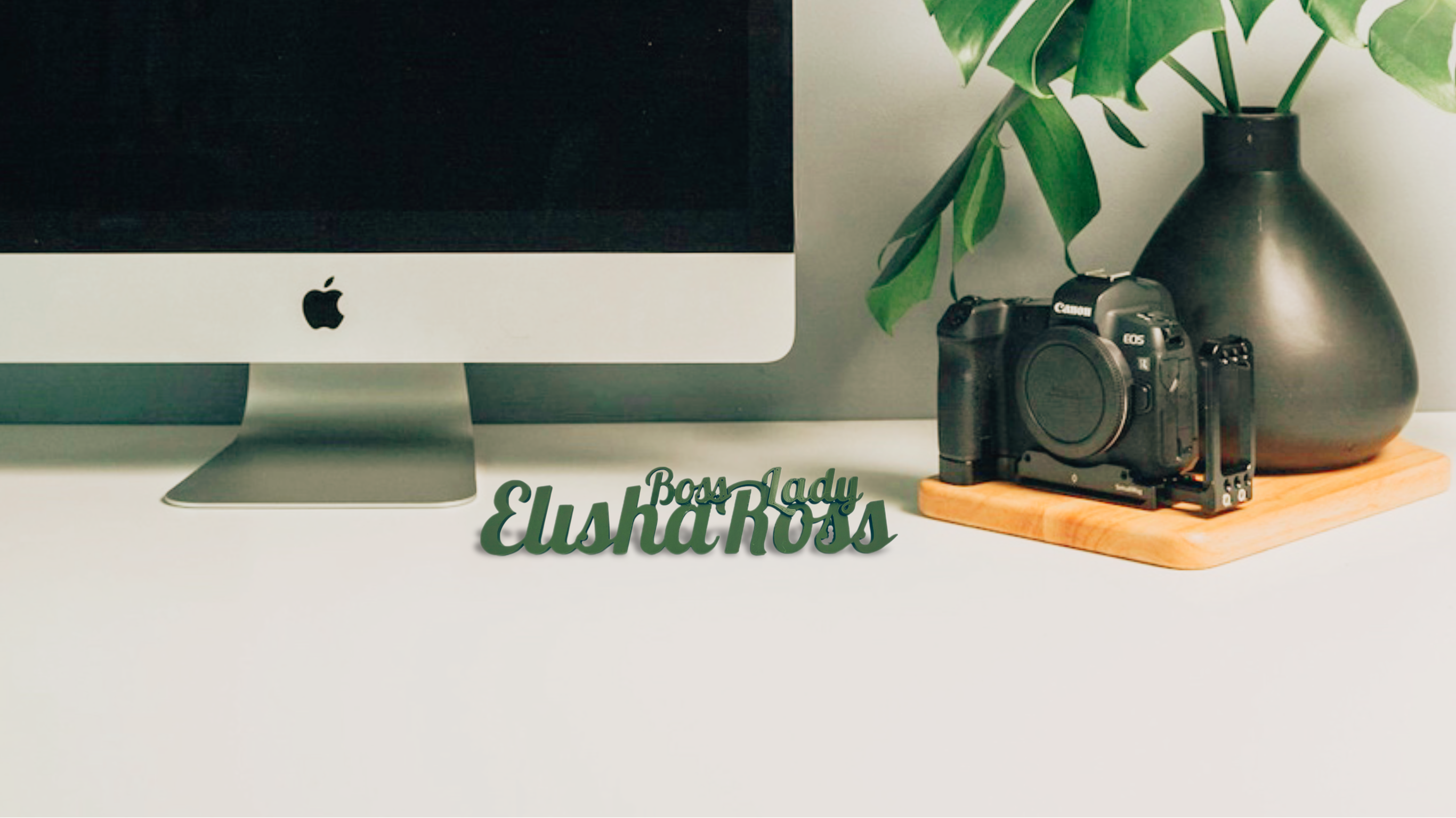 A green 3D-printed desk nameplate with "Boss Lady Elisha Ross" in a script font is positioned on a white desk. In the background, to the left, is the lower portion of an iMac computer with the Apple logo visible. To the right, a black DSLR camera sits on a wooden board next to a dark grey vase holding a large green monstera leaf. The background wall is a light grey.