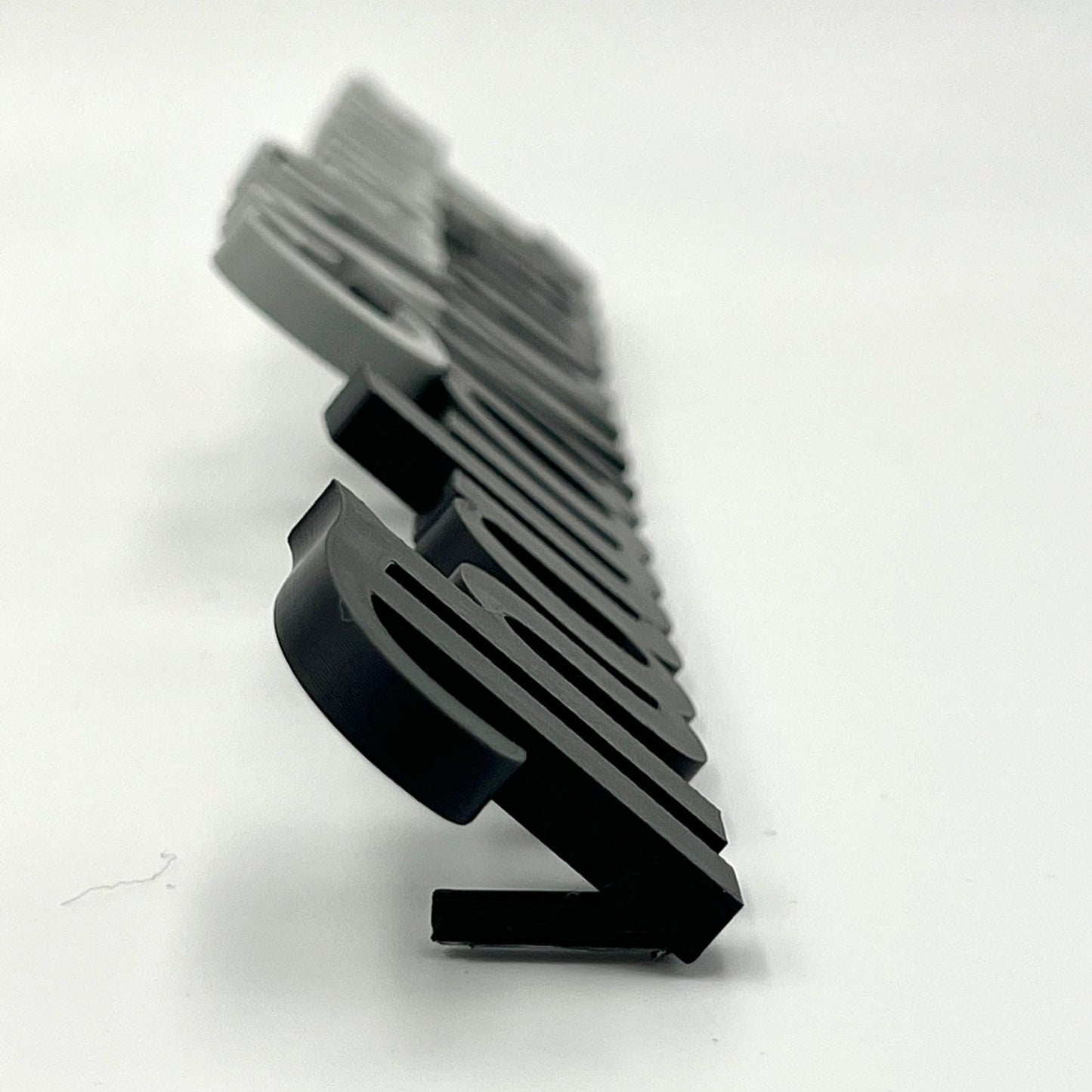 A close-up side view of several 3D-printed desk names in black and grey, showing their depth and the small stands that allow them to sit upright.

