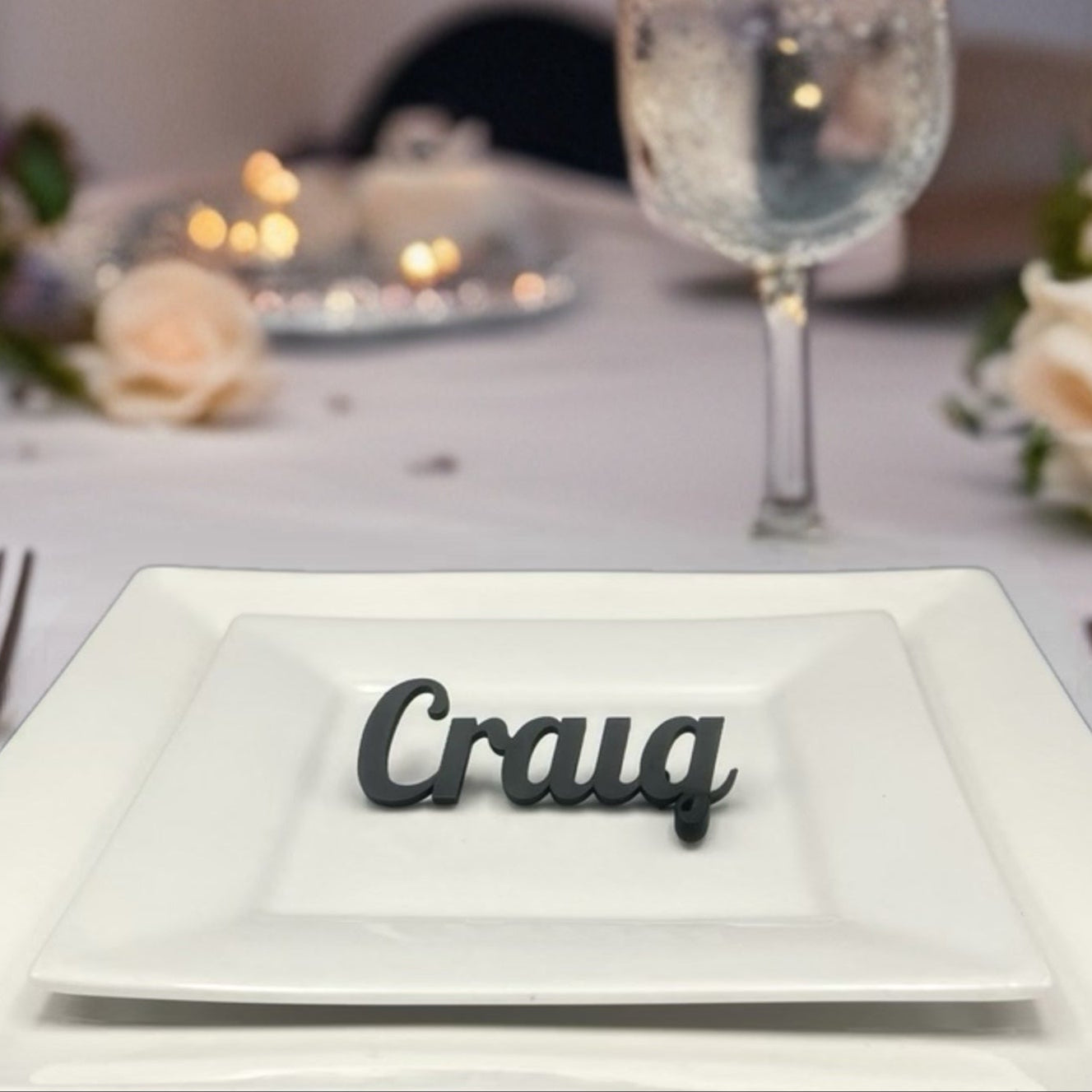 A close-up shot shows a white square plate on a white tablecloth, with a black 3D-printed name card on the plate that reads "Craig". There's a fork to the left and a knife to the right of the plate. In the background, there's a floral arrangement and a glass of water with a blue rim.