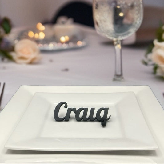 A close-up shot shows a white square plate on a white tablecloth, with a black 3D-printed name card on the plate that reads "Craig". There's a fork to the left and a knife to the right of the plate. In the background, there's a floral arrangement and a glass of water with a blue rim.