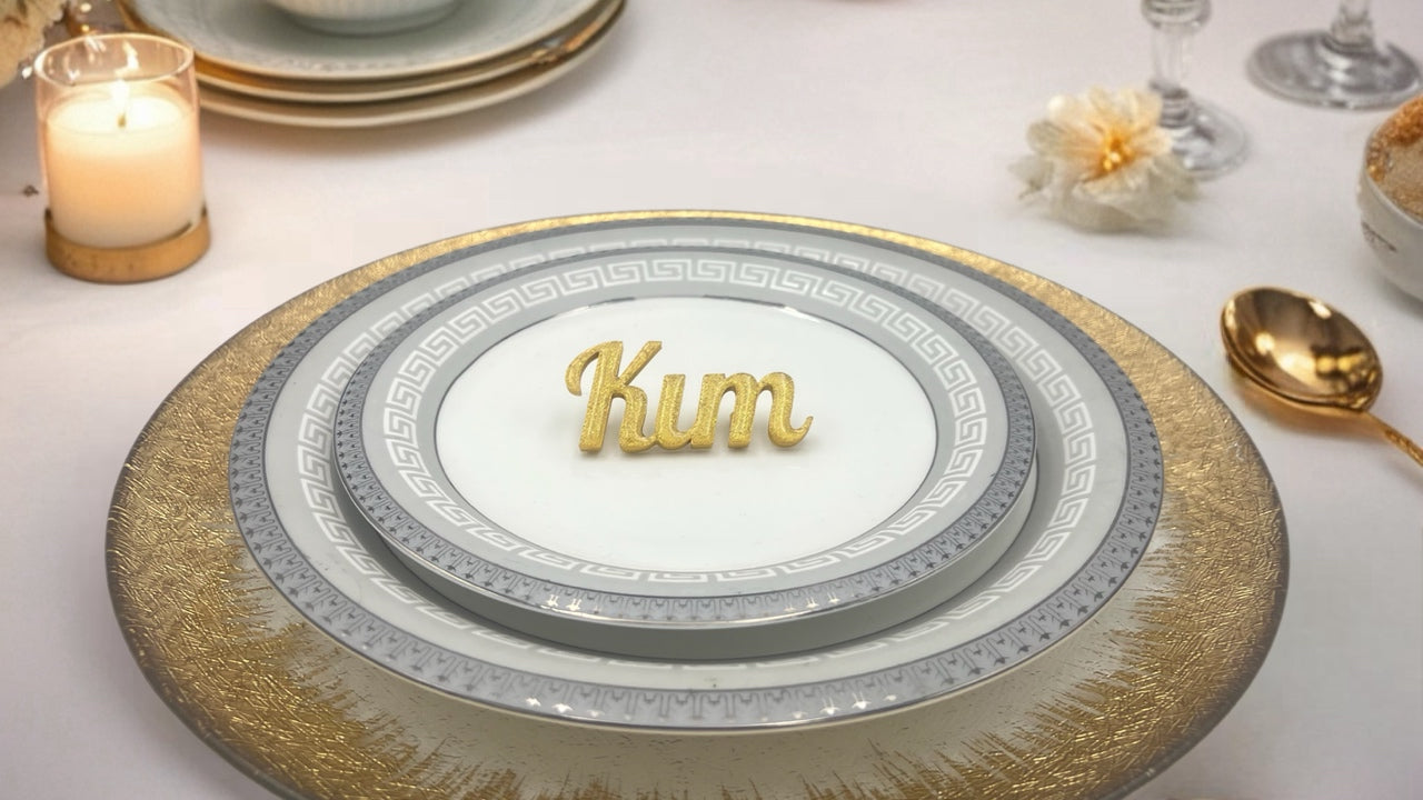 A close-up shot of an elegant wedding table setting features a stack of white and grey plates with a gold Greek key pattern, resting on a textured gold charger plate. A custom 3D-printed gold name card with the name "Kim" in a script font is placed on the top plate. A lit candle in a glass holder and a gold spoon are visible on the white tablecloth beside the plates.